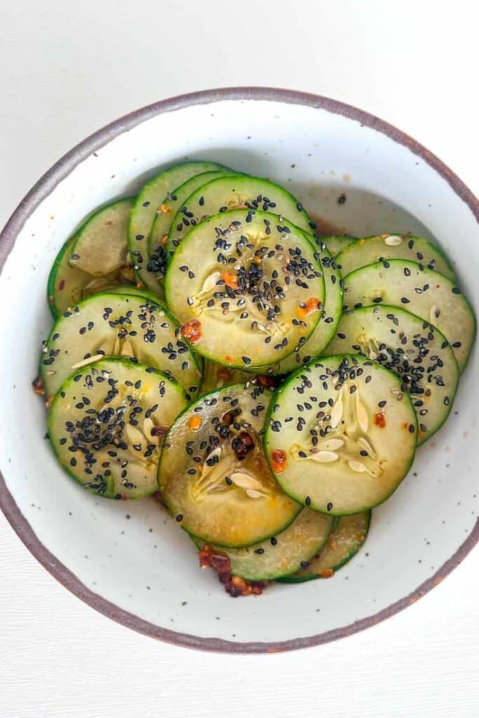 Sliced cucumbers seasoned with black sesame seeds and spices in a white bowl, showcasing a fresh and flavorful salad.