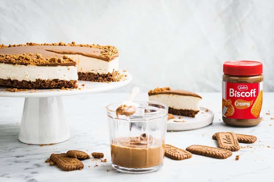 Biscoff cheesecake on a stand with a jar of Biscoff spread and cookies, on a marble countertop.