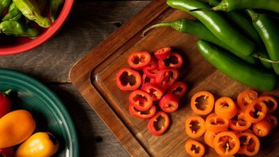 Assorted sliced bell peppers and green chilies on a wooden cutting board with colorful bowls nearby.