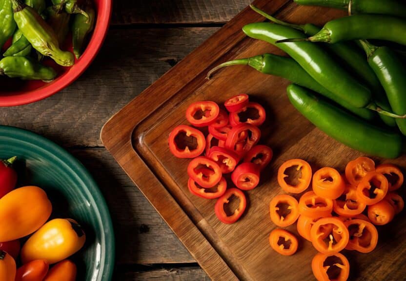 Assorted sliced bell peppers and green chilies on a wooden cutting board with colorful bowls nearby.