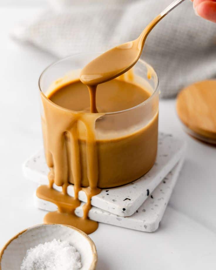 Creamy caramel sauce in a glass jar with spoon, set on marble coaster, sprinkled with sea salt, creating a sweet treat.
