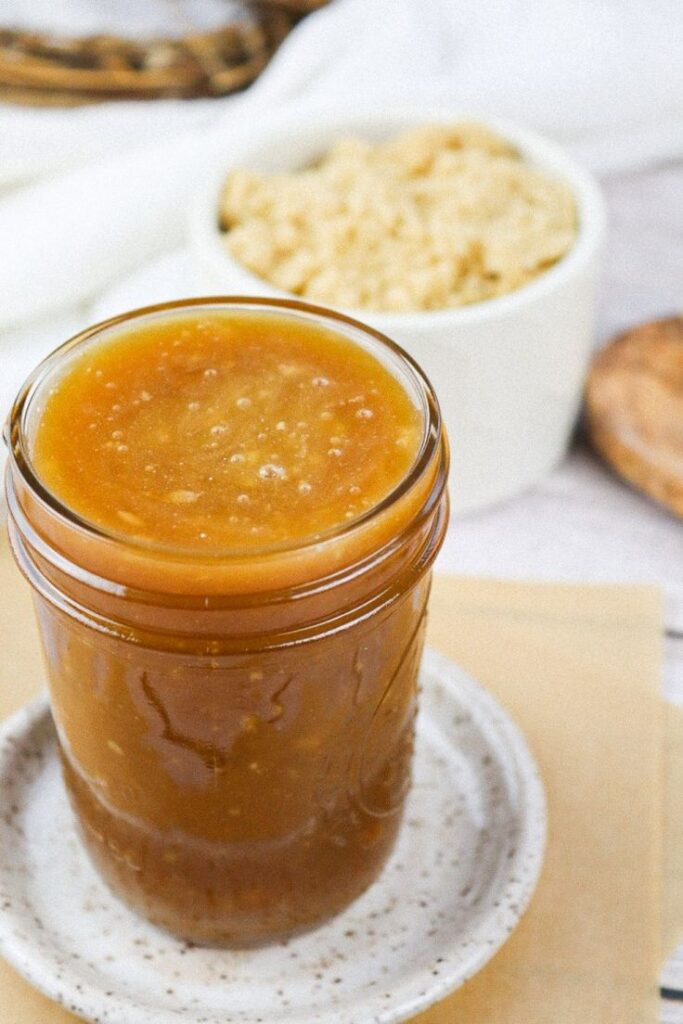 Homemade caramel sauce in a glass jar on a speckled plate with rice krispies in the background.
