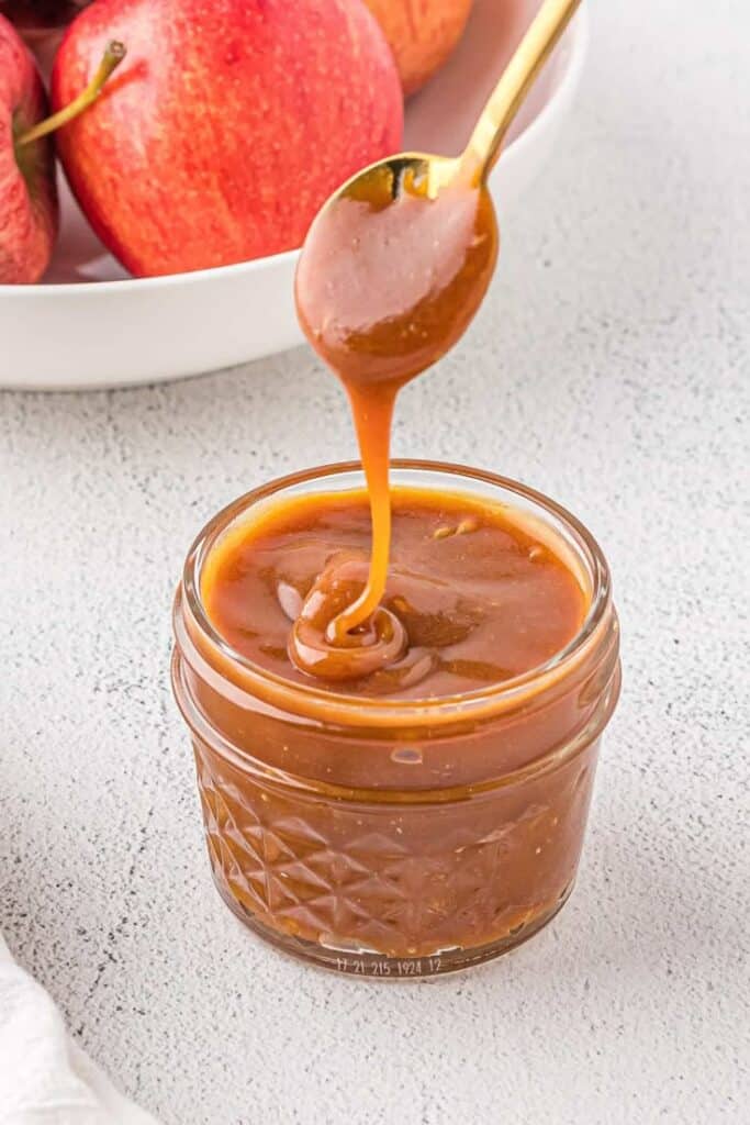 Rich caramel sauce dripping from a spoon into a jar, with fresh apples in the background on a light surface.