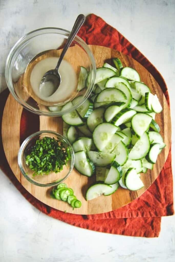 Sliced cucumbers, herbs, and dressing on a wooden board with red cloth for a fresh salad preparation.