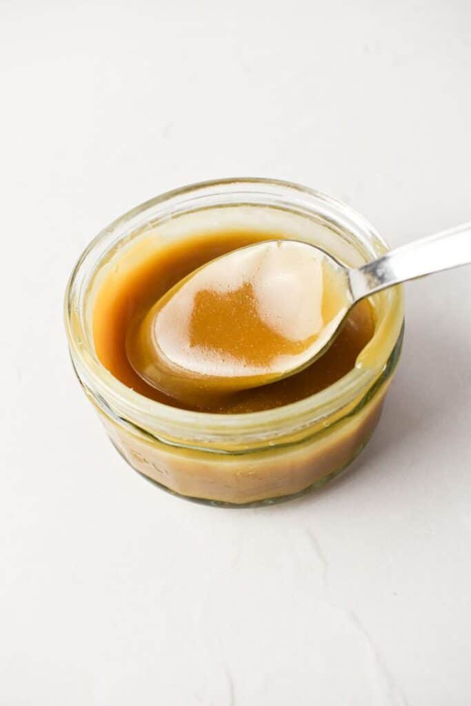 Spoon dipping into a jar of creamy caramel sauce on a white background.
