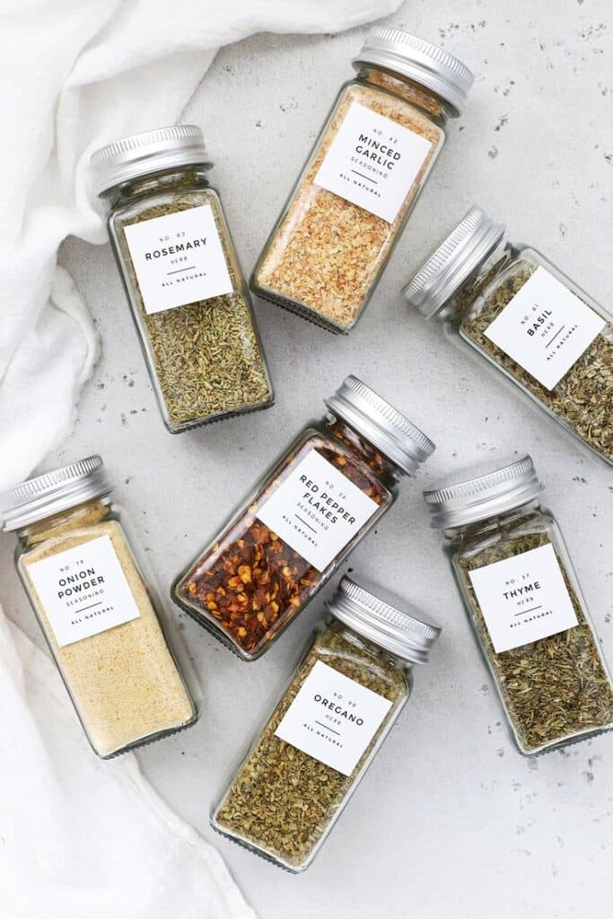 Assorted spice jars on a light countertop, including rosemary, garlic, and red pepper flakes.
