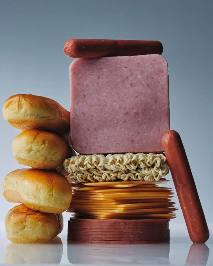 A quirky stack of buns, sausages, ham, noodles, and cheese against a blue background.