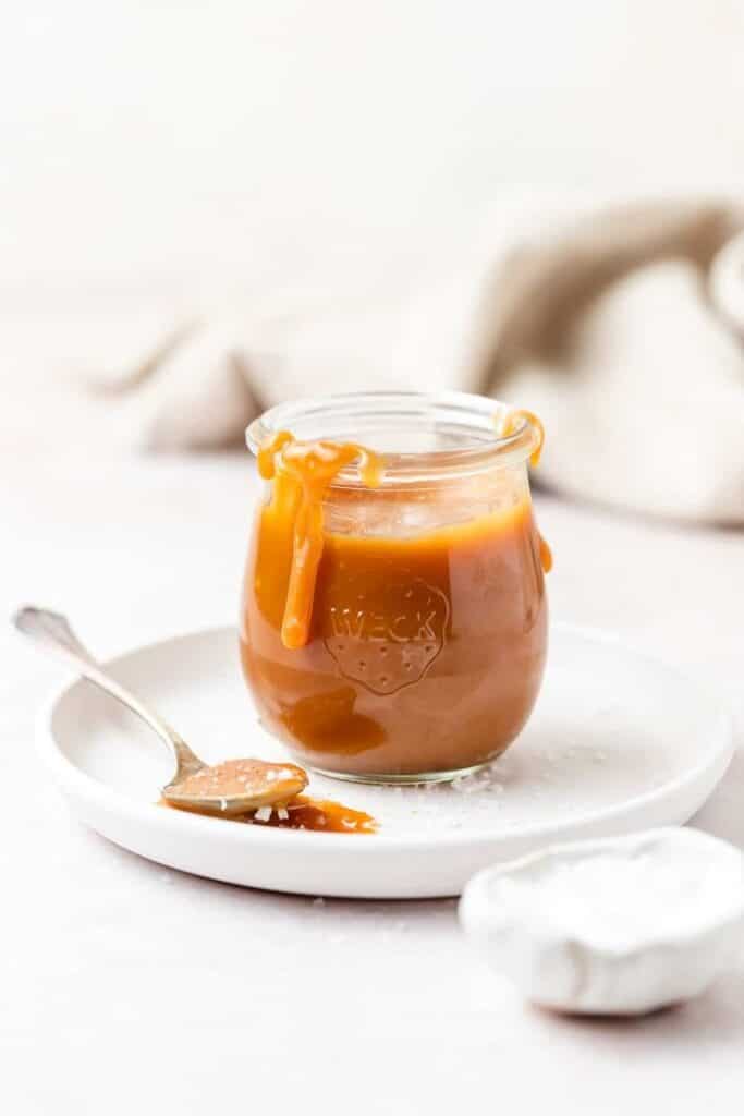 Glass jar of homemade caramel sauce with a spoon on a white plate.
