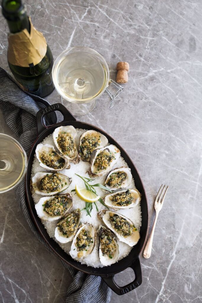 Baked oysters with herbs served with lemon and champagne on a marble table.