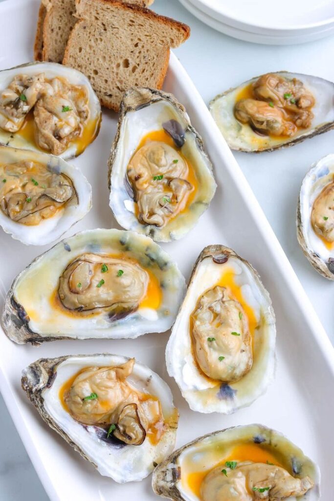 Fresh oysters served on the half shell with lemon butter sauce and bread slices on a white platter.