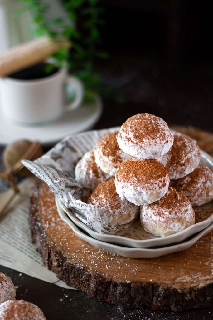 Plate of powdered cookies dusted with cocoa on a rustic wooden surface, accompanied by a steaming cup of coffee.