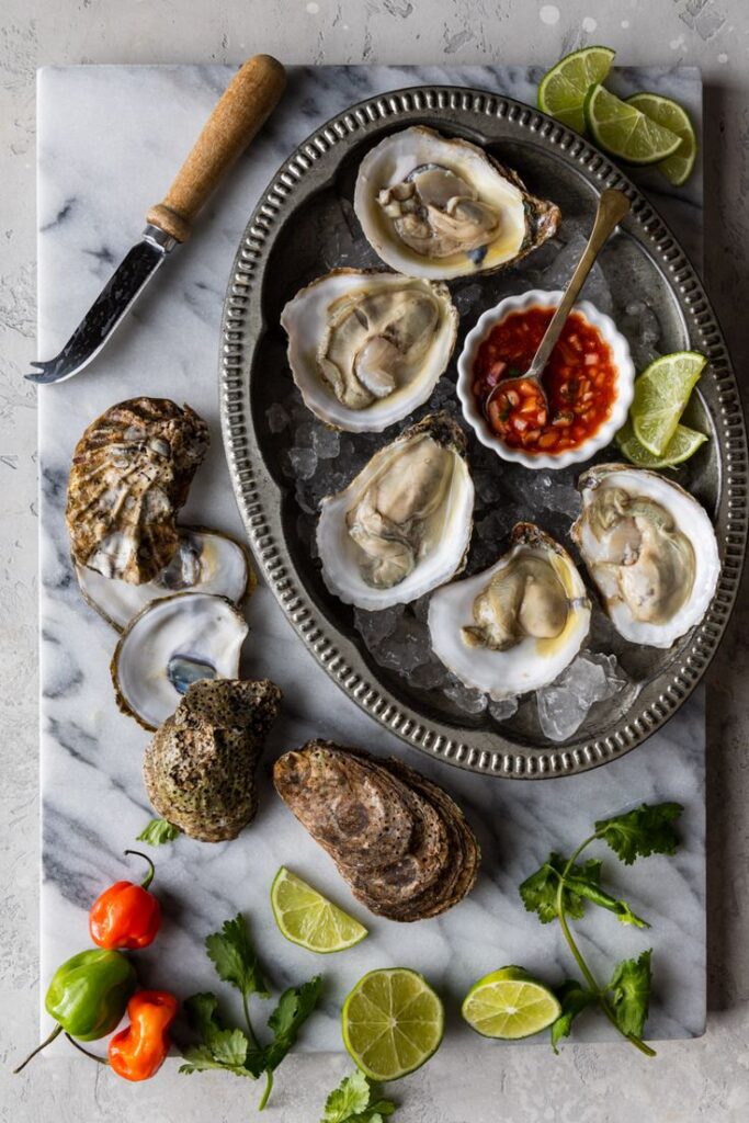 Plate of fresh oysters on ice with lime, chili peppers, and sauce on a marble surface.