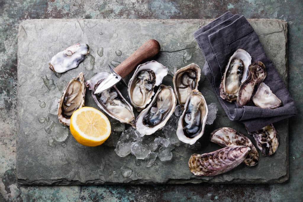 Freshly shucked oysters on ice with a lemon wedge and oyster knife on a stone slab.