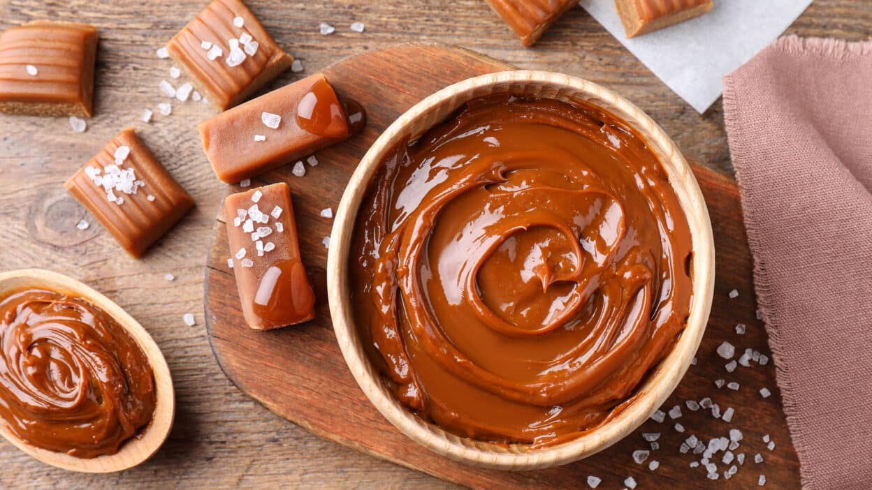 Creamy caramel sauce in a bowl with caramel candies and sea salt on a wooden table. Delicious dessert ingredients.