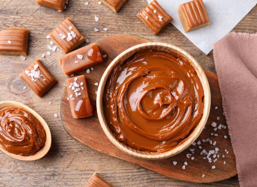 Creamy caramel sauce in a bowl with caramel candies and sea salt on a wooden table. Delicious dessert ingredients.