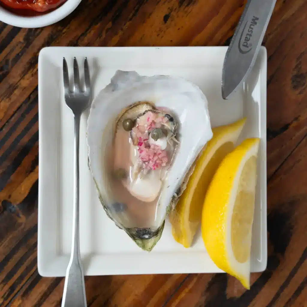 Fresh oyster on a plate with lemon wedges and sauce, served with a fork, on a rustic wooden table.