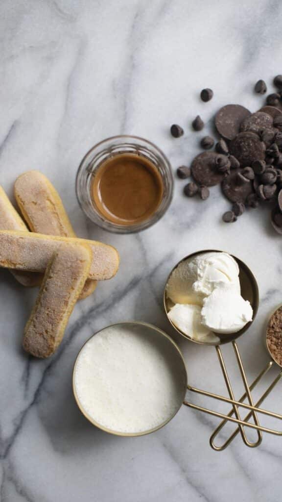 Tiramisu ingredients on marble: ladyfingers, coffee, chocolate chips, mascarpone, cocoa, cream for dessert preparation.
