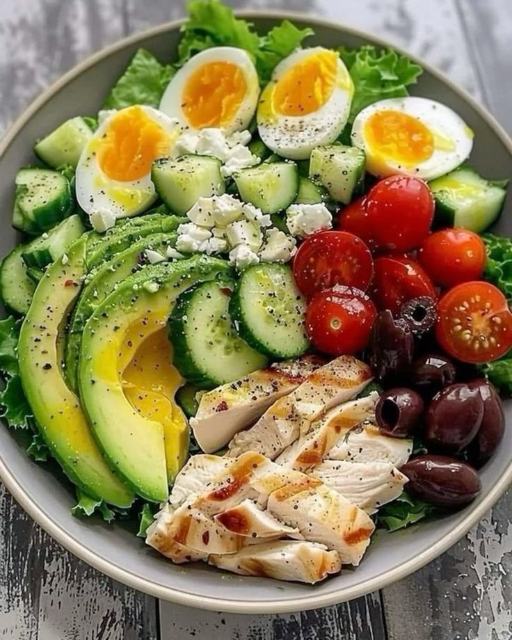 A colorful salad bowl with avocado, cucumber, eggs, tomatoes, grilled chicken, olives, and feta on a bed of lettuce.