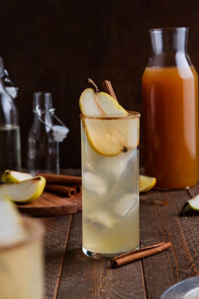 Tall glass of pear cocktail with cinnamon sticks, surrounded by fresh ingredients on a rustic table.