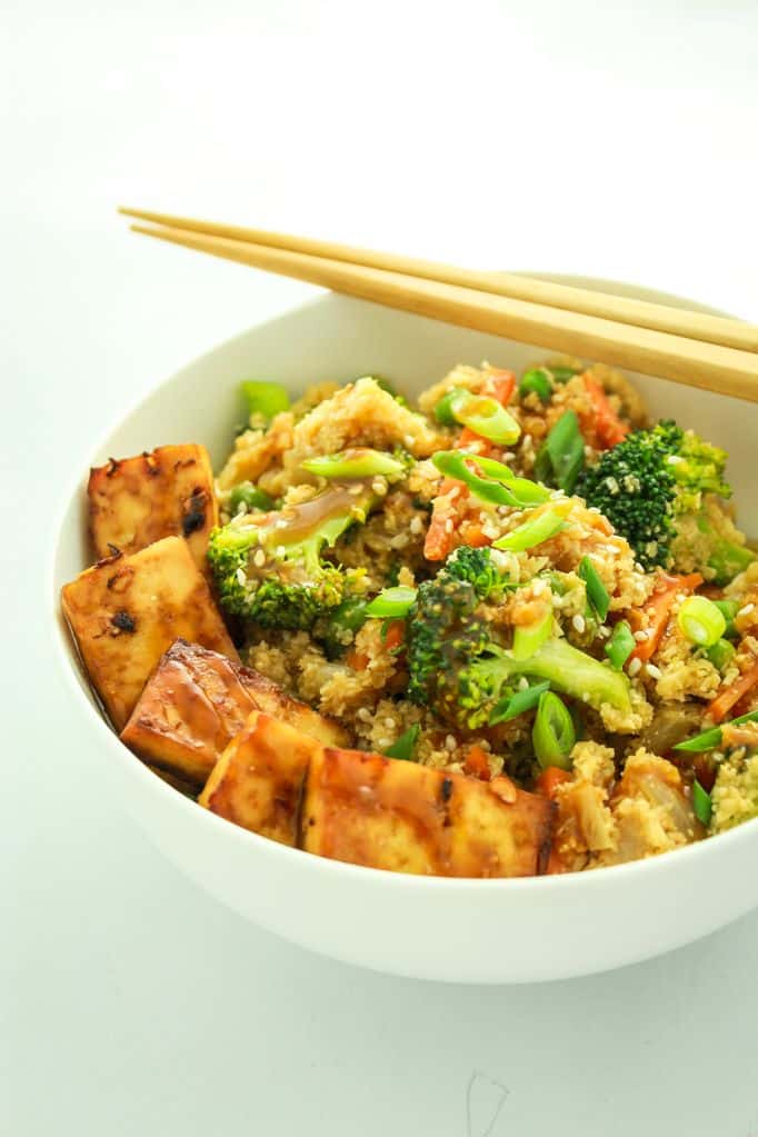 Bowl of colorful vegetable quinoa with grilled tofu and chopsticks on a white background.
