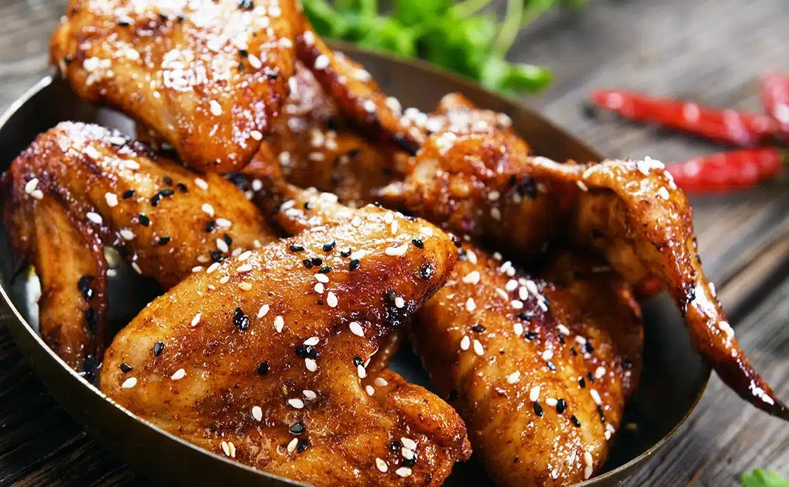 Glazed chicken wings with sesame seeds on a wooden table, garnished with herbs and red peppers.