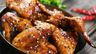 Glazed chicken wings with sesame seeds on a wooden table, garnished with herbs and red peppers.
