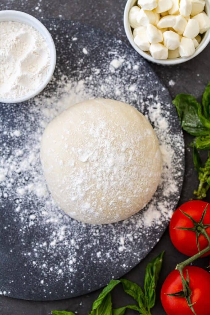 26a6cc45-08c3-427c-a443-cae7741ec653 - Master the Art of Homemade Pizza with Fun, Simple Steps | Pizza dough ball with flour, mozzarella, tomatoes, and basil on a dark surface. Perfect for making homemade pizza.