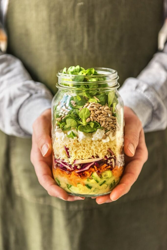 Layered salad in a jar with couscous, greens, and seeds held by a person in an apron.