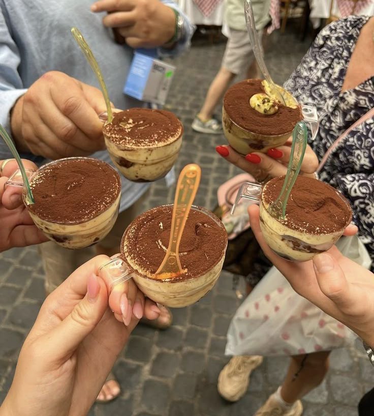 People holding cups of tiramisu with cocoa topping, celebrating together outdoors.