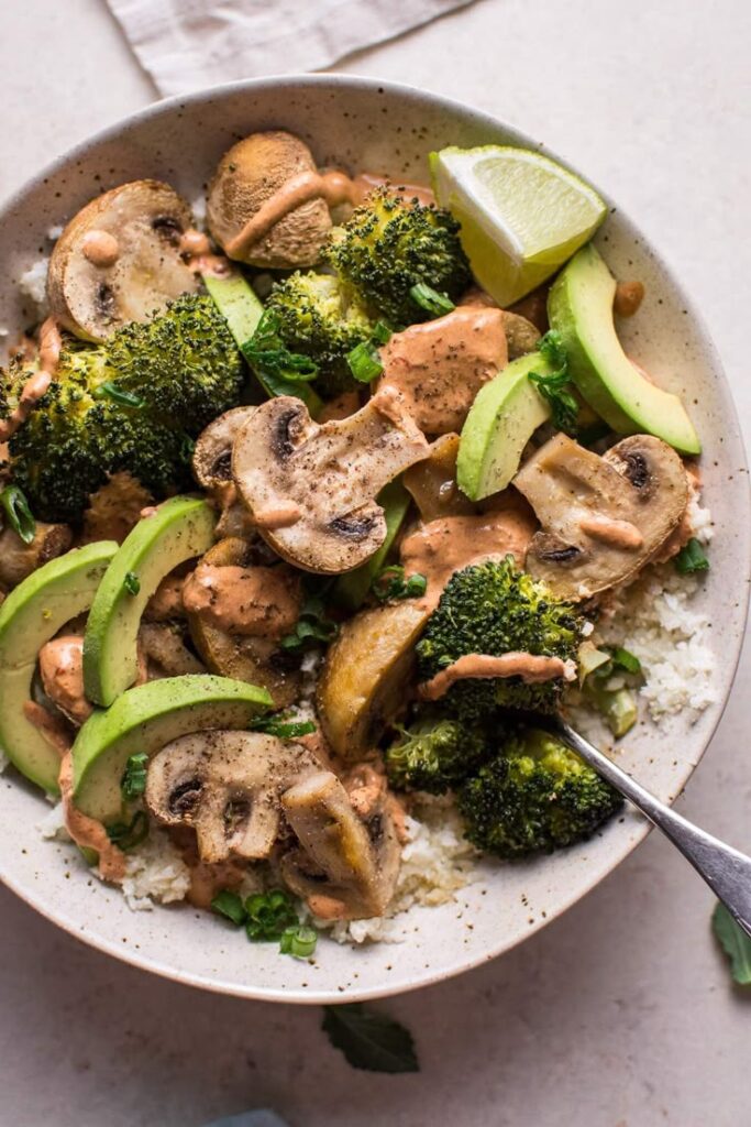 Healthy bowl with broccoli, mushrooms, avocado, and sauce on a bed of rice, garnished with lime wedge.