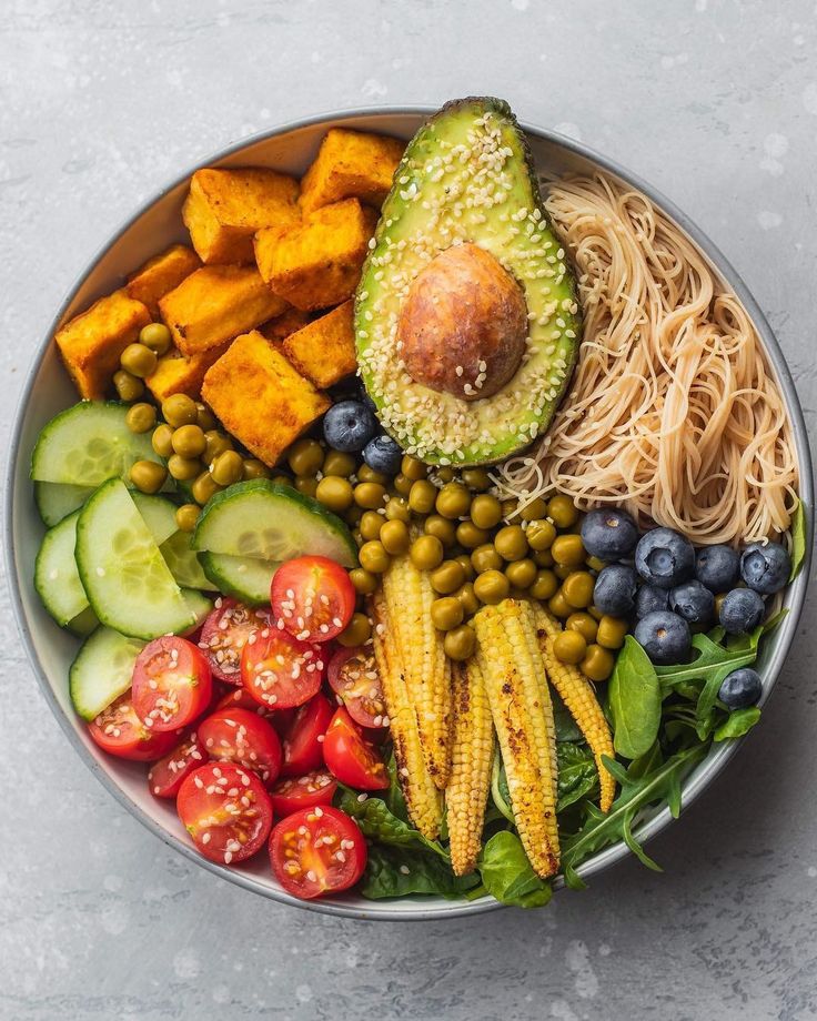 Colorful vegan Buddha bowl with avocado, sweet potato, noodles, cherry tomatoes, blueberries, and baby corn on a gray background.