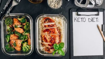 Keto meal prep containers with grilled chicken, broccoli, and rice next to a notepad labeled Keto Diet.