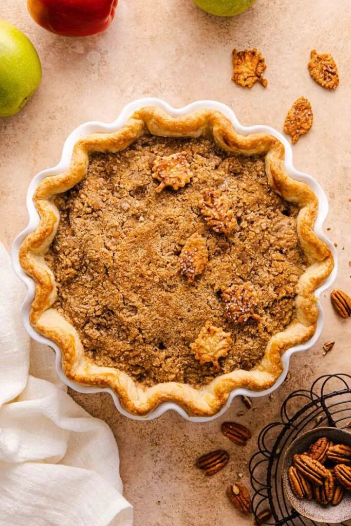 Homemade apple pie with streusel topping in a fluted dish, surrounded by ingredients on a rustic table.