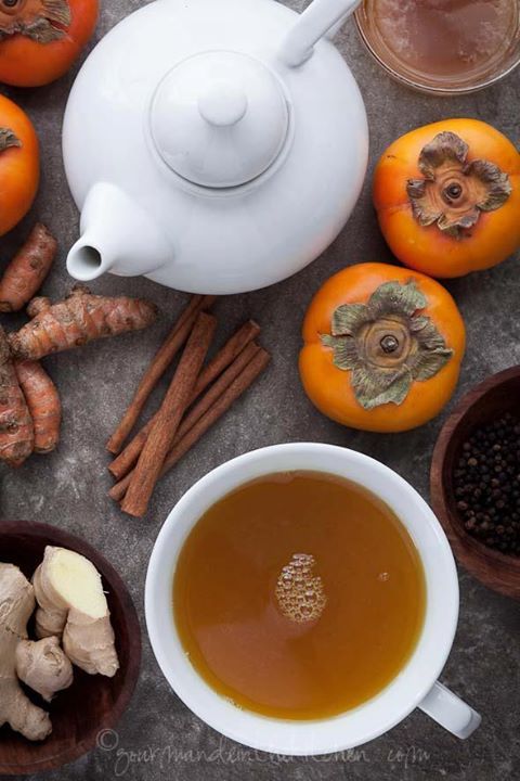 Tea and ingredients flat lay with white teapot, fresh persimmons, ginger, turmeric, cinnamon sticks, and black pepper.
