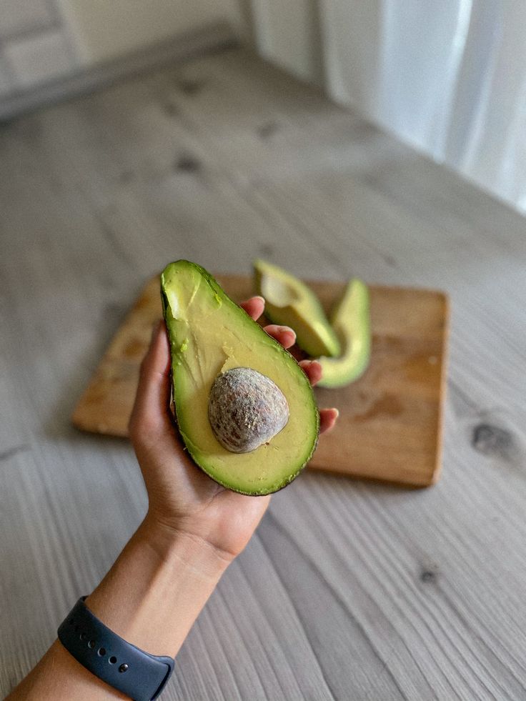 Hand holding a ripe avocado half with pit over wooden board, slices in background. Fresh, healthy ingredient.