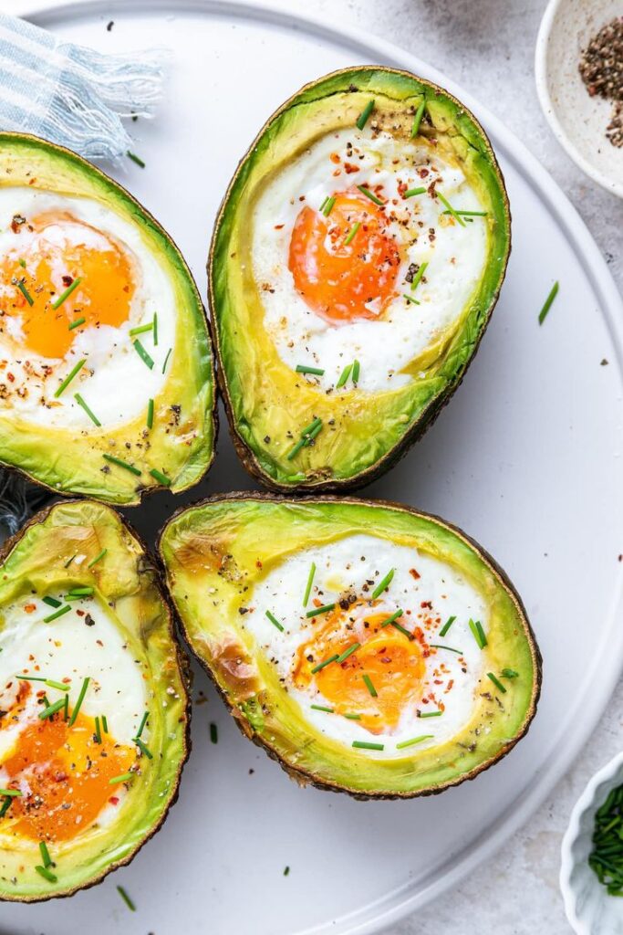 Avocado halves with baked eggs, garnished with chives and pepper, on a white plate for a healthy breakfast idea.