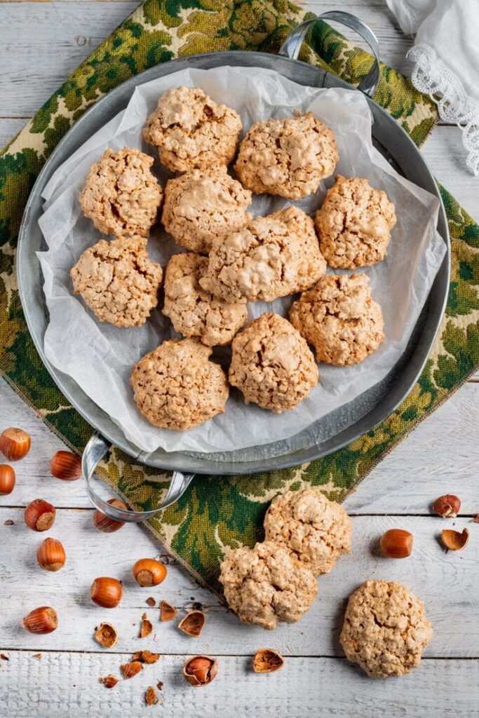 Rustic homemade hazelnut cookies on parchment paper in a metal tray, surrounded by hazelnuts on a wooden table.