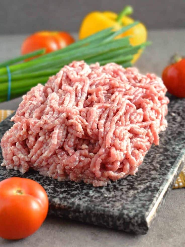 Fresh ground meat on a stone slab with tomatoes, bell pepper, and green onions in the background.