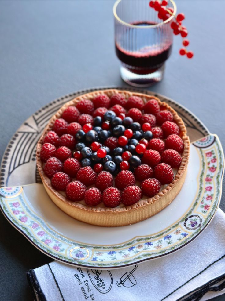 Berry tart with raspberries, blueberries, and currants on a decorative plate beside a glass of red juice.