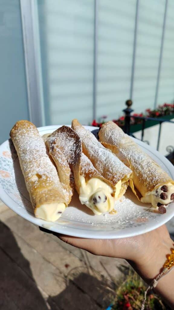 Plate of cannoli with powdered sugar and creamy filling, set against a sunny outdoor background, held by a hand.