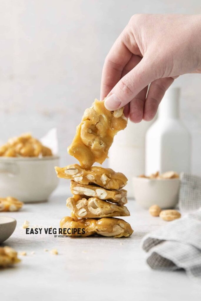 Hand holding a peanut brittle piece above a stack, with bowls and fabric in the background.