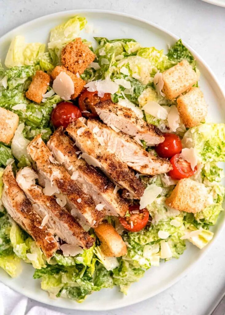Grilled chicken Caesar salad with cherry tomatoes, croutons, and parmesan on a white plate.