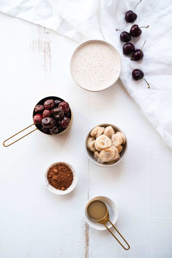Top view of smoothie ingredients: cherries, bananas, cacao powder, nut milk, and syrup on a white surface.