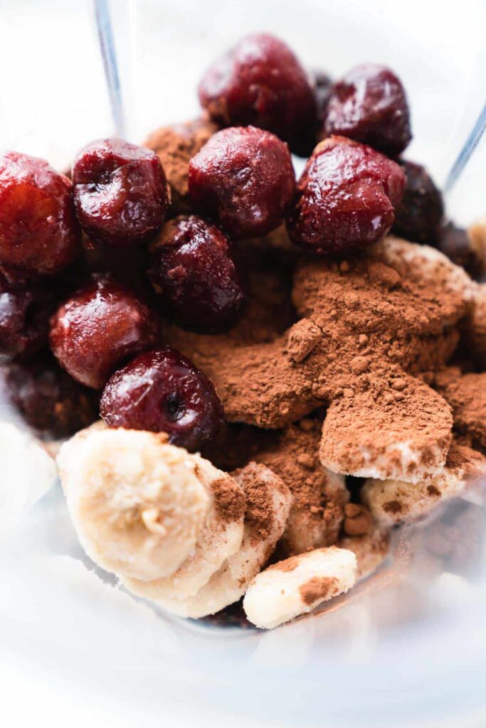Frozen cherries, banana slices, and cocoa powder for a smoothie in a blender.