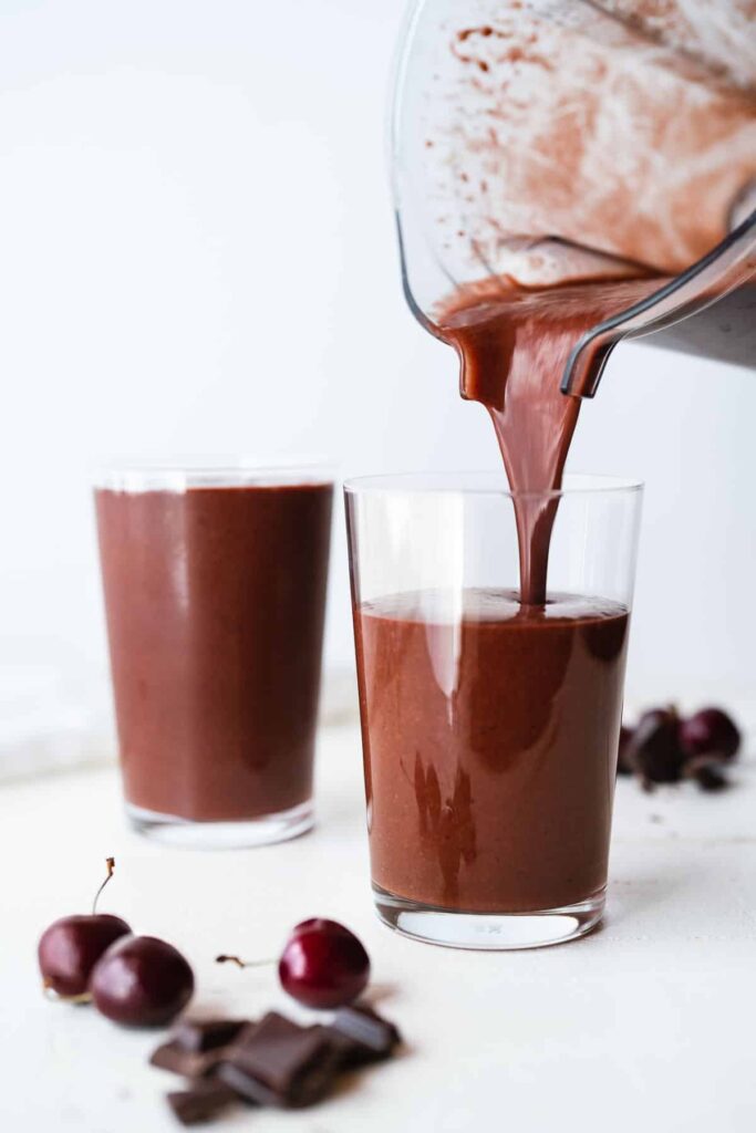 Chocolate cherry smoothie pouring into a glass, with fresh cherries and chocolate pieces nearby.