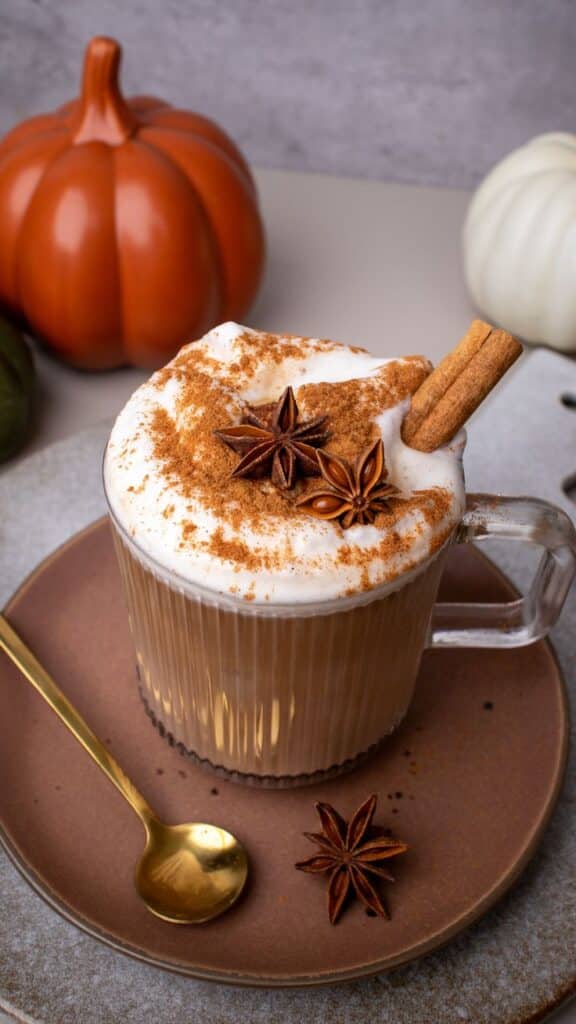 Festive spiced latte with frothy cream, cinnamon, and star anise, surrounded by pumpkins on a plate with a golden spoon.