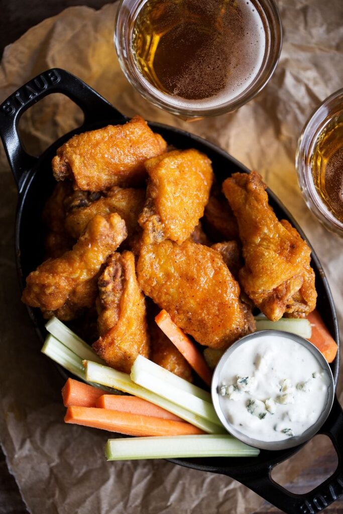 Close-up of crispy buffalo wings with celery, carrots, and blue cheese dip in a black dish beside glasses of beer.