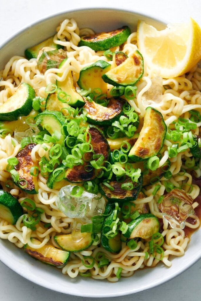 Bowl of noodles with zucchini, green onions, and lemon slice, garnished with ice cubes for a refreshing twist.