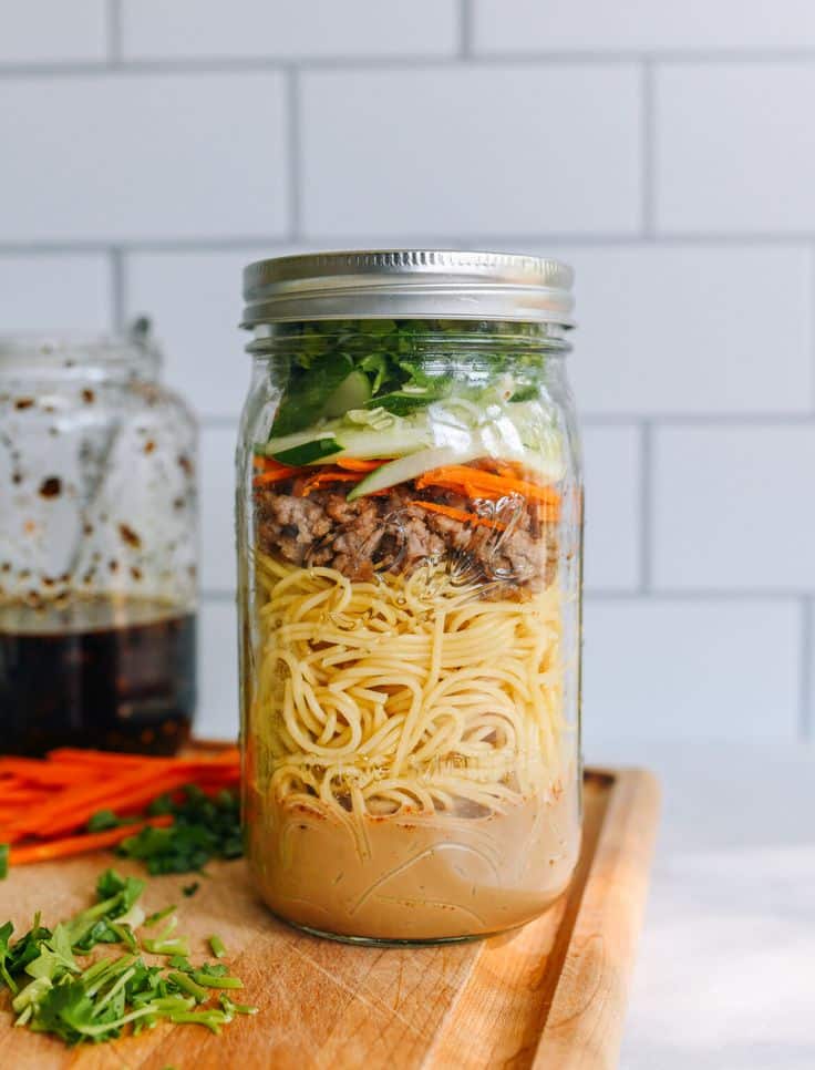 Layered noodle jar with vegetables, meat, and sauce on a wooden board in front of a white tile wall.