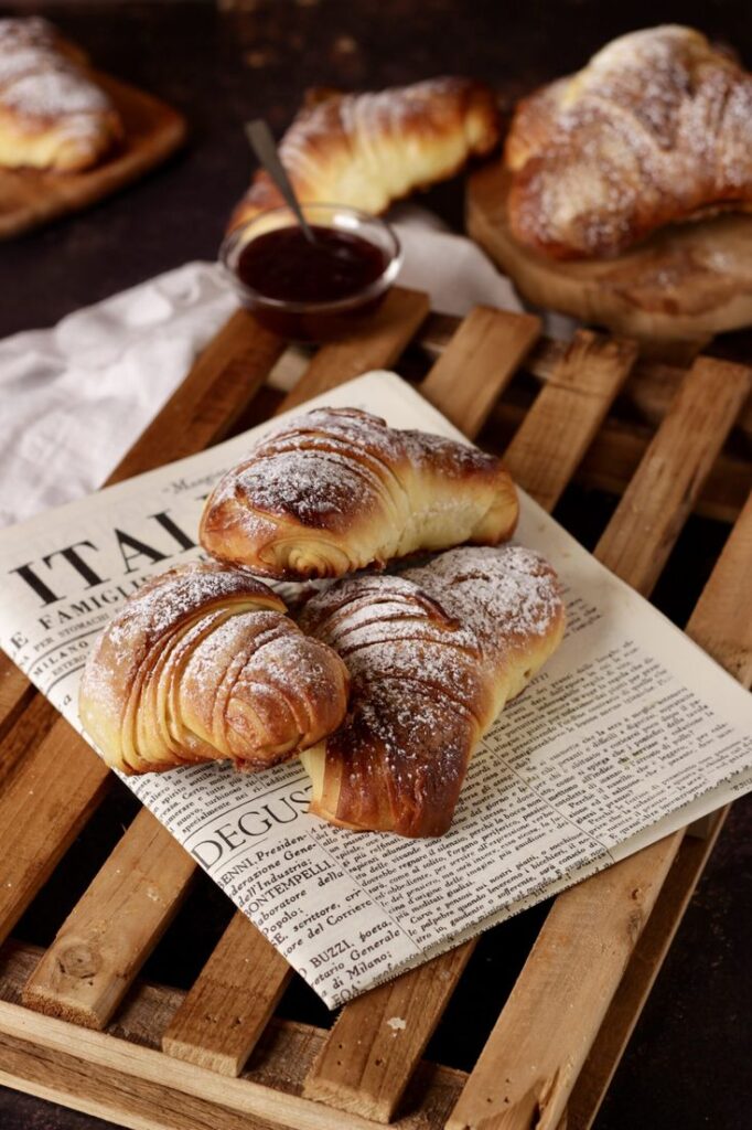 Flaky croissants sprinkled with powdered sugar on a wooden board, accompanied by a small jar of jam.